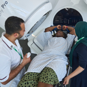 A person in a hospital gown is laying down. They have their arms above their head in holsters. On either side of them are a healthcare professional. One is operating a radiotherapy machine that is focused on the person laying down's chest.